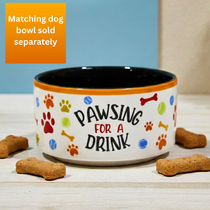 Dog bowl with 'Pawsing for a Drink' text and colorful paw print design on a wooden surface with dog treats. Sold separately from a matching mug.
