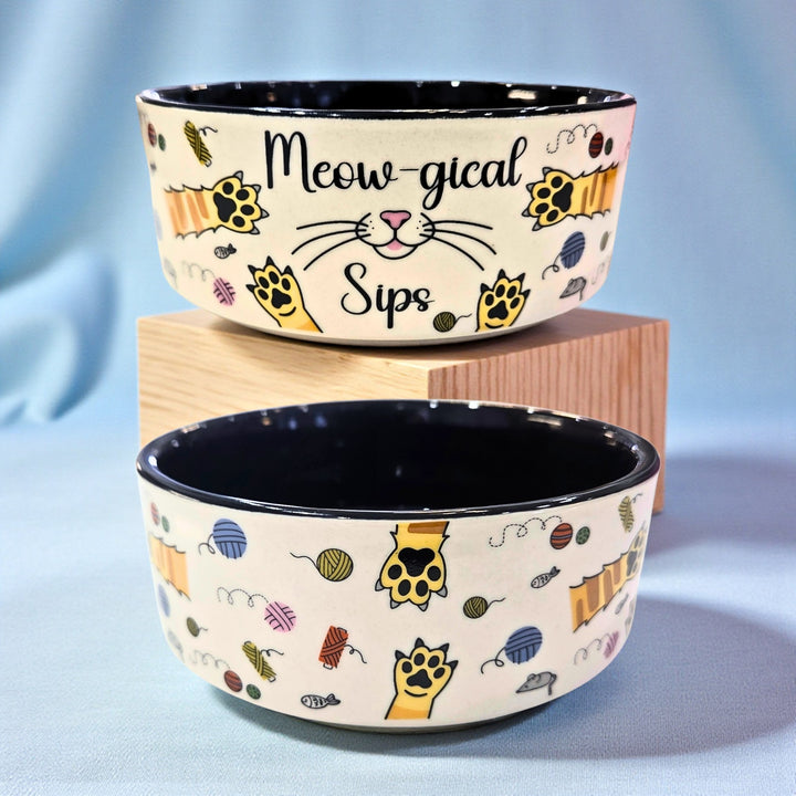Front and side view of a cat-themed pet bowl with text saying "Meowgical Sips" on one side and illustrations of yarn, cat paws, and mice around it.