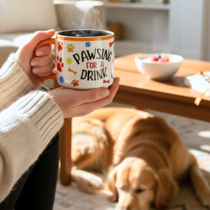 Person holding a mug with 'Pawsing for a Drink' text, dog lying on the floor in the background